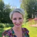 Kvinna ler i soligt utomhuslandskap med grön bakgrund.