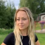 En kvinna med långt blont hår står ute i en trädgård, iklädd svart skjorta, tar en paus från sina städningsplikter; träd och en byggnad utgör bakgrunden.