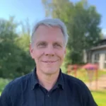 En leende man med grått hår framför en grönskande bakgrund.