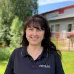 Kvinna med mörkt hår och en svart pikétröja som ler utomhus framför en byggnad och träd.