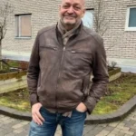 Man klädd i brun skinnjacka och jeans står utanför en tegelbyggnad och ler — kanske uppskattar den pålitliga fastighetsservicen från SRL Fastigheter.