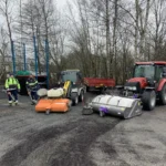 Två arbetare står vid fordon med stora gatusopare och förbereder grusupptagning under vårstädning på en gård med kala träd i bakgrunden.