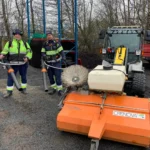 Två arbetare i säkerhetsutrustning står vid ett gatsoparfordon med orange Crenova-utrustning, upptagna med grusupptagning — en riktig vårtecken utomhus.