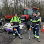 Två arbetare i synlig utrustning står med lövblåsare, redo för grusupptagning och vårstädning, framför en röd traktor och sopmaskin.